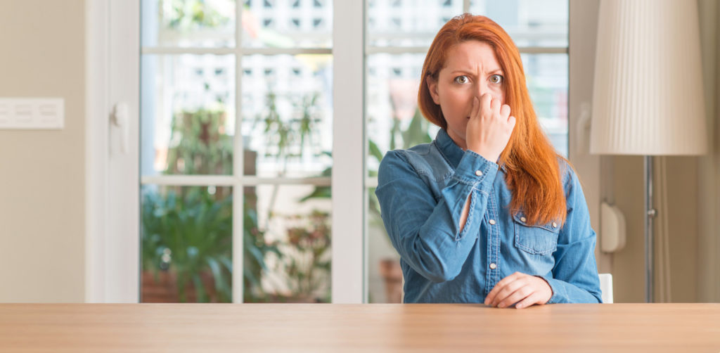Redhead woman at home smelling something stinky and disgusting, intolerable smell, holding breath with fingers on nose.