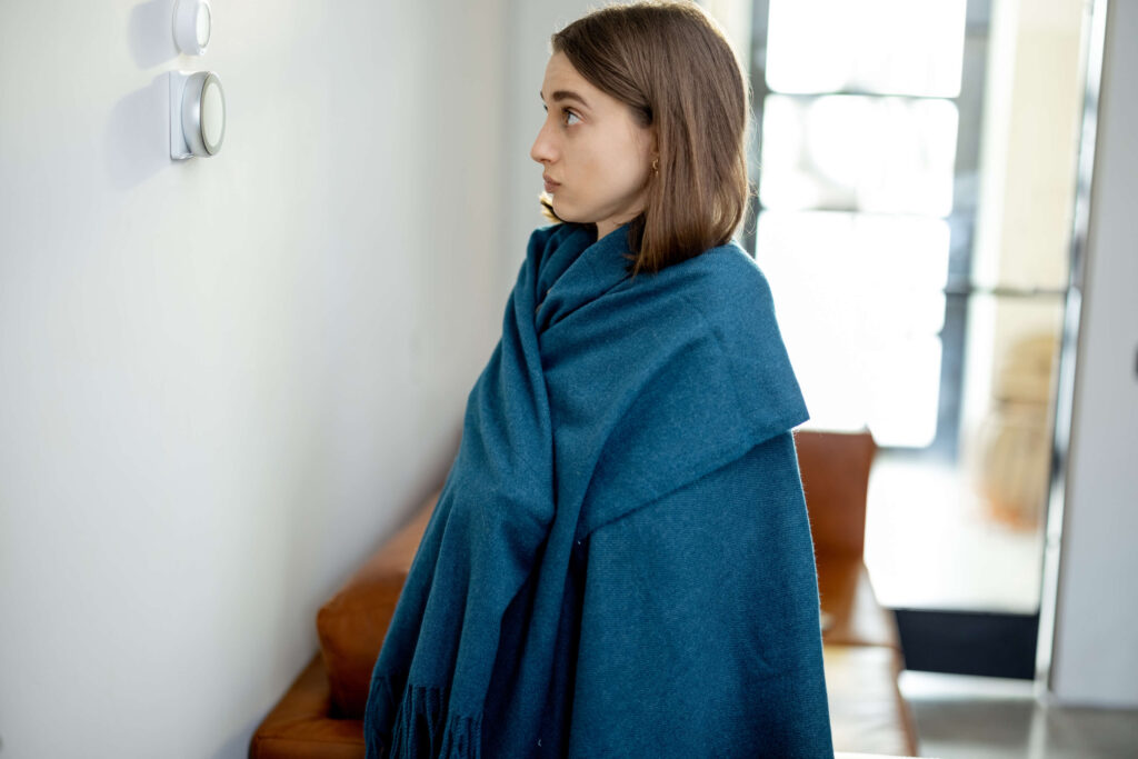 woman wrapped in blue blanket looking at wall thermostat not turning on heater